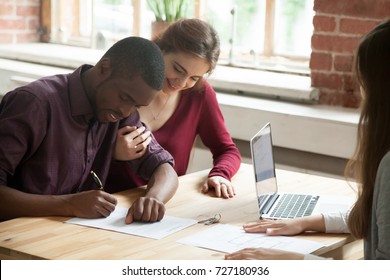 African american man signing home purchase agreement in front of realtor, his smiling wife watching contract being signed. Young multiethnic family leasing new apartment. First time buyers concept. 