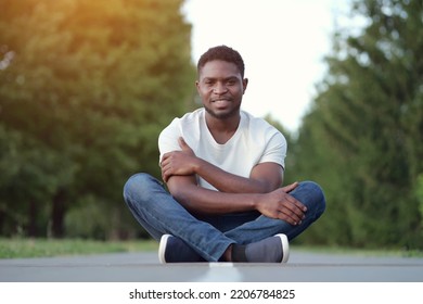 African American Man Sitting Cross Legged On Asphalt Shows Toothy Smile And Enjoys Life. Young Guy Sits Against Blurry Trees In Park At Twilight