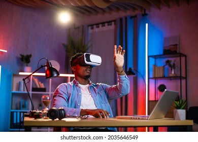 African american man sitting at home office and using VR glasses and laptop for remote work. Black male freelancer touching virtual screen with fingers. - Shutterstock ID 2035646549