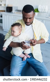 African American Man Sitting On Couch With Baby And Messaging On Smartphone