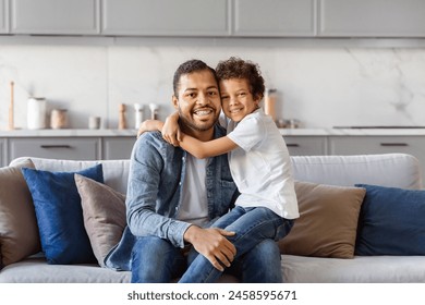 African American man is sitting on top of a couch next to a little boy. The father appears relaxed, while the son looks curious. They seem to be engaged in a conversation or activity together. - Powered by Shutterstock