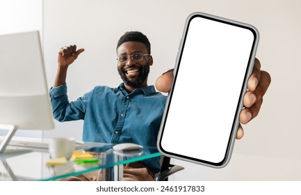 African American man sitting at workdesk with pc, holding a cell phone in his hand, showing blank white screen and gesturing - Powered by Shutterstock