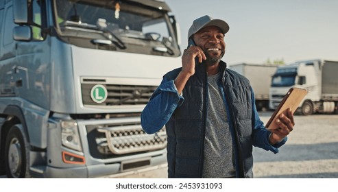 African American man speaking on phone with his coworker or customer while holding tablet device. Busy worker standing at trucks parking while remotely communicating. Concept of technology. - Powered by Shutterstock