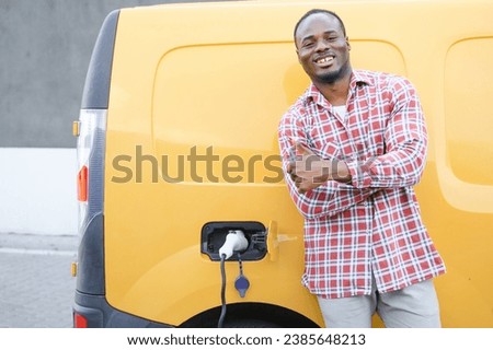 A african american man stands next to yellow electric delivery van at electric vehicle charging stations.