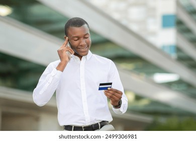 African American man stands outdoors, smiling as he examines his credit card while conversing on his mobile phone. He is engaged, possibly discussing financial matters or making a purchase decision - Powered by Shutterstock