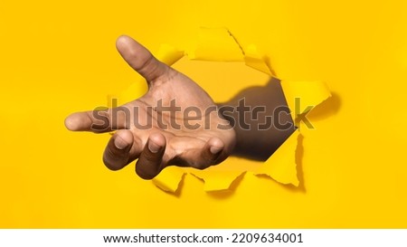 African american man stretching open palm through hole in torn yellow paper, offering helping hand, taking or giving something, asking for charity, reaching out for support