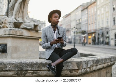 African American Man In Stylish Suit And Hat Sitting On Old Fountain And Holding Modern Smartphone. Handsome Young Guy Surfing Internet Among City Street.