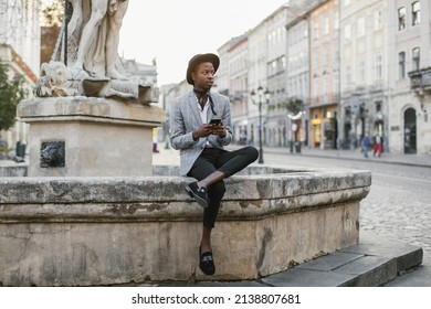 African American Man In Stylish Suit And Hat Sitting On Old Fountain And Holding Modern Smartphone. Handsome Young Guy Surfing Internet Among City Street.