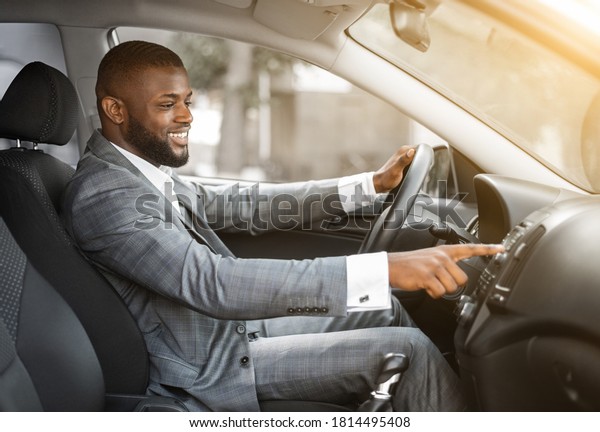 African american man in suit driver turning on music\
in car before moving. Cheerful black businessman pushing button on\
functional panel in his car, turning on music before starting\
business trip