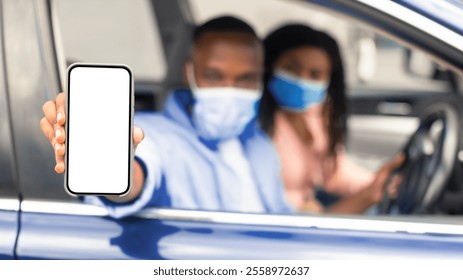 African American man in surgical mask showing mobile phone with white empty screen for mock up sitting on driver seat in car with woman, holding gadget in hand, selective focus on device - Powered by Shutterstock