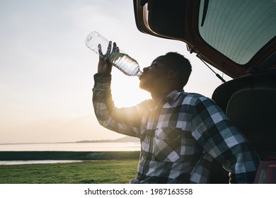 African American Man Tourists Relax In Front Of Their Cars And Drink Thirsty Water From Bottle With Sunlight. In Evening Atmosphere Of The Dam With Mountains And Good Weather For Relaxing And Camping