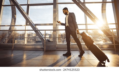 An African American man is using his phone in an airport terminal, standing with his luggage. The setting is modern and bustling, indicating efficient travel, sun flare - Powered by Shutterstock