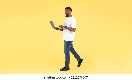 African American Man Using Laptop Computer Walking Working Online On The Go Posing Over Yellow Studio Background. Internet Technology And Freelance Career. Panorama, Full Length Shot