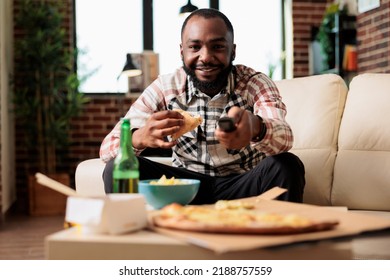 African American Man Using Tv Remote Contorl To Switch Channel And Eating Slice Of Pizza From Fast Food Takeout Delivery. Having Fun With Takeaway Meal And Beer While He Watches Movie.
