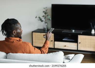 African American Man Watching Television, Turning On Plasma Flatscreen TV-set, Pointing Remote Control At Empty TV Screen, Mockup. Black Guy Switching Channels At Home, Back View