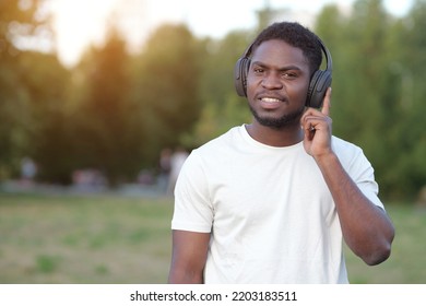 African American Man Wearing Black Headphones Listens To Favourite Music. Black Guy Enjoys Walking In Park Against Blurry Green Trees