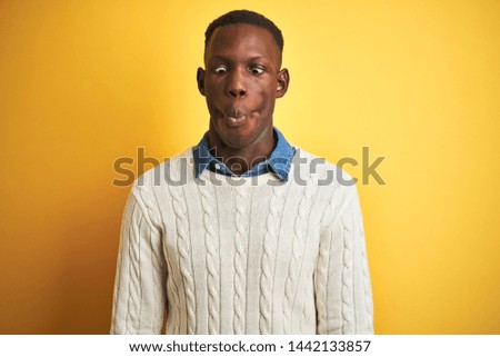 African american man wearing denim shirt and white sweater over isolated yellow background making fish face with lips, crazy and comical gesture. Funny expression.