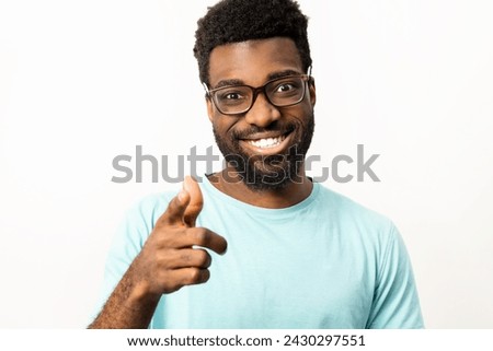 African American man wearing glasses and a casual t-shirt smiling and pointing towards the camera on an isolated white background, representing positivity and friendliness.