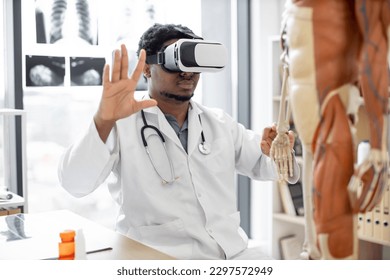 African american man wearing lab coat and 3d goggles using VR system while sitting near modern devices and human skeleton in office. Therapist preparing for medical training in teaching hospital. - Powered by Shutterstock