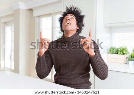 African American man wearing winter sweater amazed and surprised looking up and pointing with fingers and raised arms.