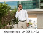 African American man in a white dress shirt and khaki pants stands on a city street while talking on his mobile phone. He has a smile on his face and is looking off to the side.