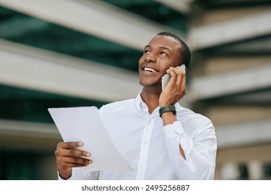 African American man in a white shirt stands outside of an office building, talking on his phone and holding a document in his left hand. He looks up with a smile and appears to be listening intently - Powered by Shutterstock