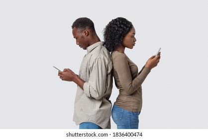 African american man and woman standing back to back, using mobile phones during quarantine, grey studio background, copy space. Black couple sitting in social media instead of talking to each other