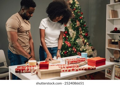 African american man and a woman wrapping Christmas gifts together on a table adorned with wrapping paper and ribbons, while a beautifully decorated Christmas tree stands in the background. - Powered by Shutterstock