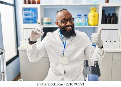 African American Man Working At Scientist Laboratory Holding Syringe Smiling Happy And Positive, Thumb Up Doing Excellent And Approval Sign 