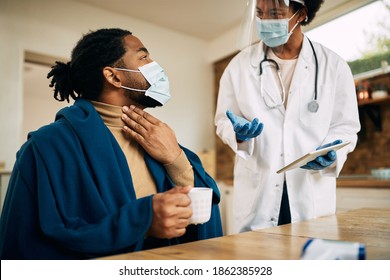 African American Man Wrapped In Blanked Talking To His Doctor And Complaining About Sore Throat During A Home Visit. Both Of Them Are Wearing Face Masks Due To Coronavirus Pandemic.