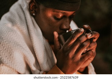 African American man wrapped in a blanket enjoys a warm drink. Black man sipping hot drink in blanket in winter. The warm drink and blanket provide comfort for black man in winter - Powered by Shutterstock