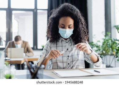 African American Manager In Medical Mask Holding Hand Sanitizer, And Businessman Working On Blurred Background