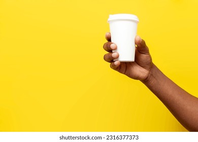 african american man's hand holds and offers white paper cup of coffee on yellow isolated background, empty package for drink in the hands of man, mock-up