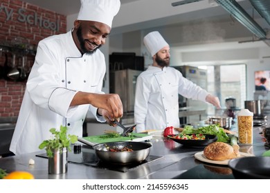African american master chef using big knife to cut organic herbs and vegetables for stew meal. Food industry worker seasoning gourmet dish prepared for dinner service at fine dining restaurant. - Shutterstock ID 2145596345