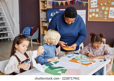 African American mature teaher helping little boy cut holes in yellow paper mask while standing by group of intercultural learners
