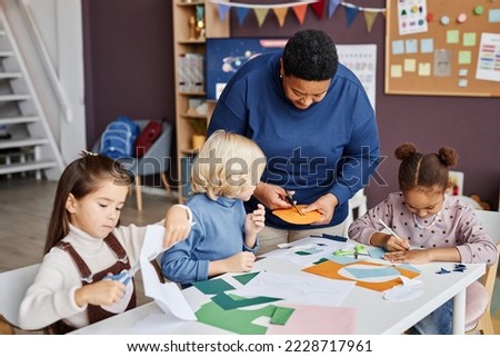 African American mature teaher helping little boy cut holes in yellow paper mask while standing by group of intercultural learners