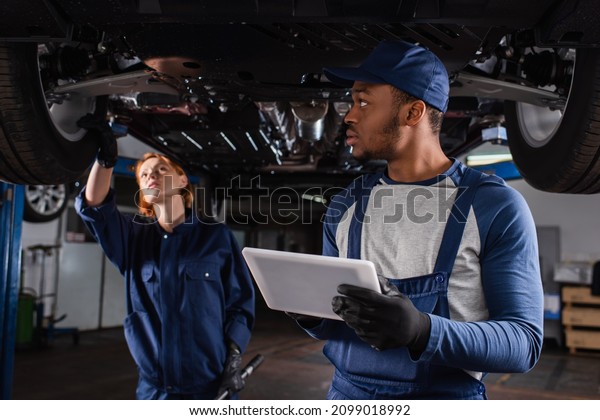 African american mechanic holding\
digital tablet while colleague standing under car in\
garage