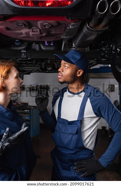 African american mechanic pointing at car bottom\
near colleague in\
garage