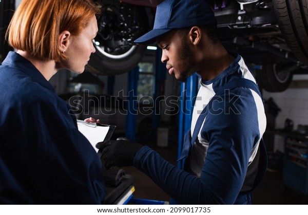 African american mechanic pointing at clipboard\
near colleague in car\
service