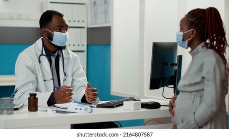 African American Medic And Patient With Pregnancy Talking About Childbirth, Doing Consultation At Office Desk. Physician And Future Mother Having Conversation During Covid 19 Pandemic.