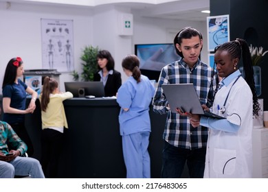 African American Medical Doctor Holding Laptop With Appointment Talking With Asian Patient In Busy Private Clinic Waiting Room. Medic Having Conversation With Young Man Checking Lab Results.