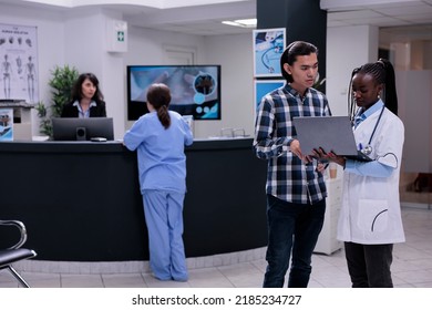 African American Medical Doctor Holding Laptop Presenting Lab Results To Asian Patient In Private Practice Clinic. Medic Talking With Young Man Checking Appointment On Portable Computer.