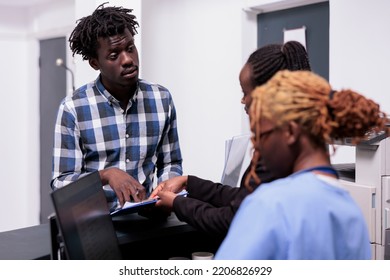 African American Medical Team Talking To Adult At Hospital Reception Desk, Filling In Report Papers Before Consultation Appointment. Patient Receiving Insurance Support From Nurse And Worker.