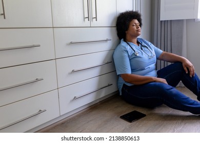 African American Mid Adult Female Nurse With Eyes Closed Sitting On Floor In Hospital. Unaltered, Healthcare Worker, Stress, Expertise And Occupation Concept.