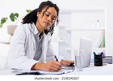 African american mid adult female doctor writing on clipboard while using laptop on desk in hospital. wireless technology, prescription, doctor, medical, healthcare, hospital and medical occupation.