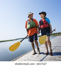 African American Middle-aged Couple Walking On Boat Dock Holding Hands And Carrying Paddles.