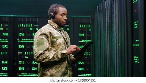 African American Military Man In Uniform And Headset Working In Servers Room, Tapping On Tablet Device And Speaking In Mic. Work With Secret Information In Army. Digital Data Defence Concept.