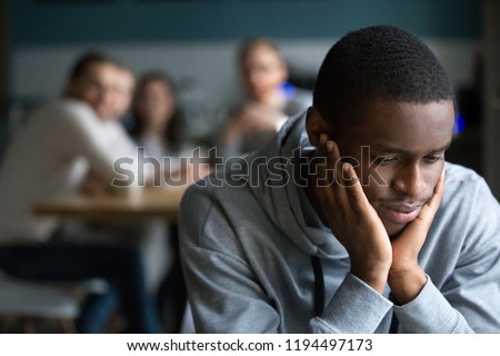 African American millennial guy feel lonely sitting alone in café, offended black student avoid talking to friends having misunderstanding, young man outcast suffer from racial discrimination