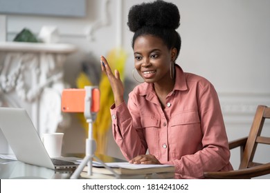 African American millennial woman with afro hairstyle remote studying, working online on laptop, chatting with friends via video call on smartphone on tripod. Blogger influencer recording video blog.
