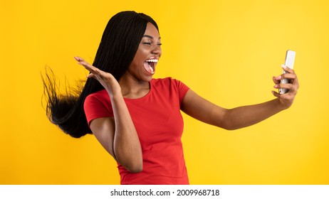 African American Millennial Woman Using Smartphone Making Selfie Playing With Braided Hair, Posing And Having Fun On Yellow Background. Studio Shot Of Excited Female Making Video Call Via Phone